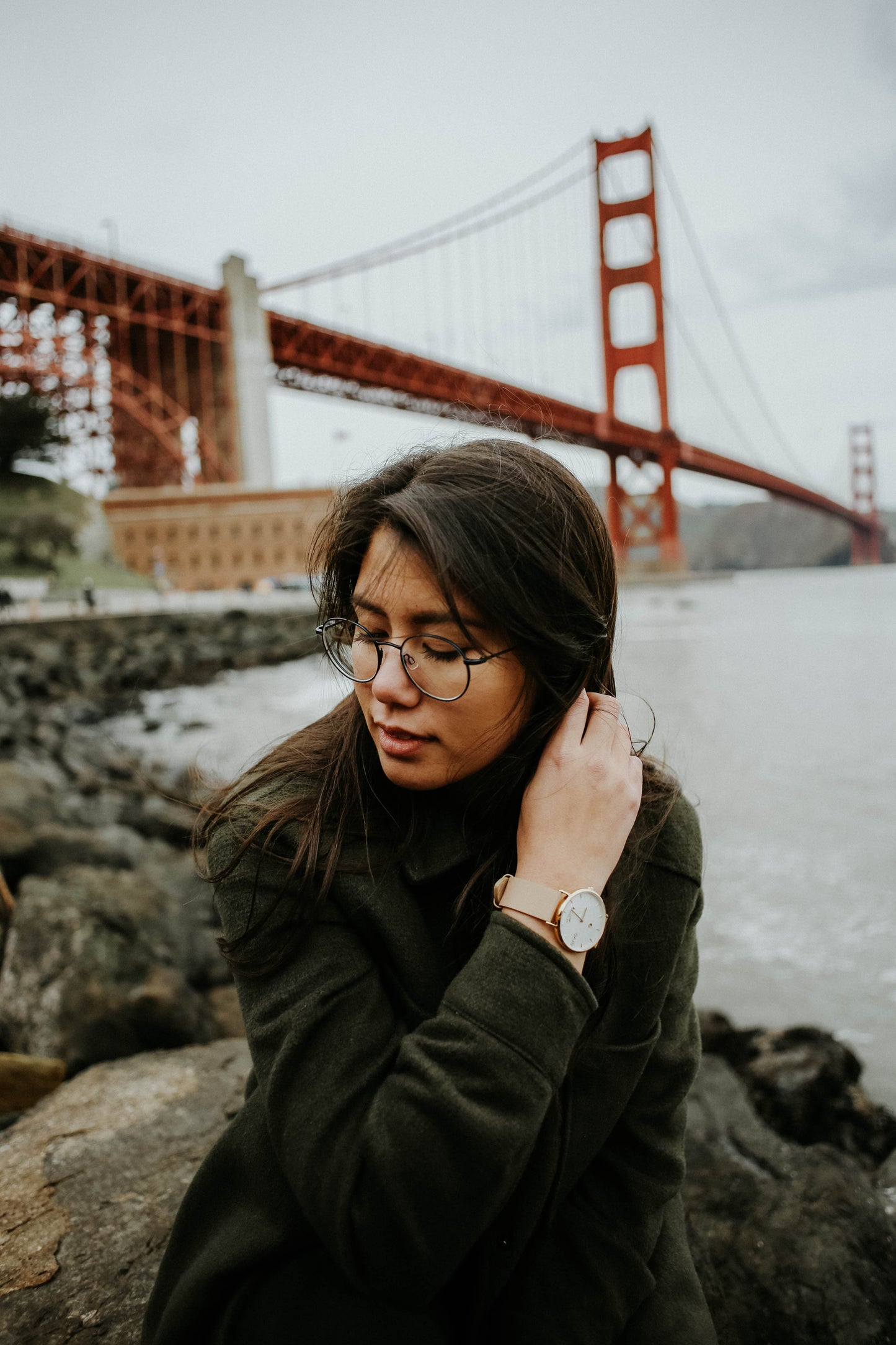 Asian woman on the shore beside the Golden Gate Bridge. She is wearing an Arvo Awristacrat gold watches for women with white dial, gold case, and nude or tan leather band