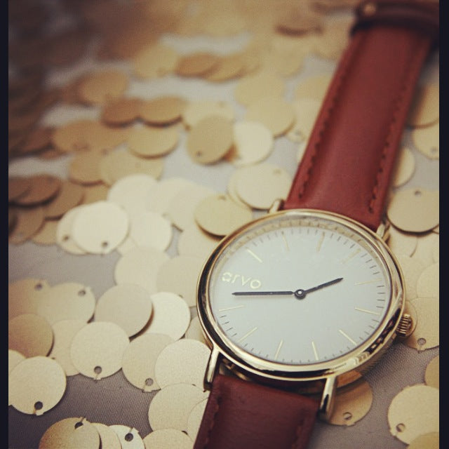 Time Sawyer watch for men and women white dial, gold case, and brown leather band laying on a bed of gold discs